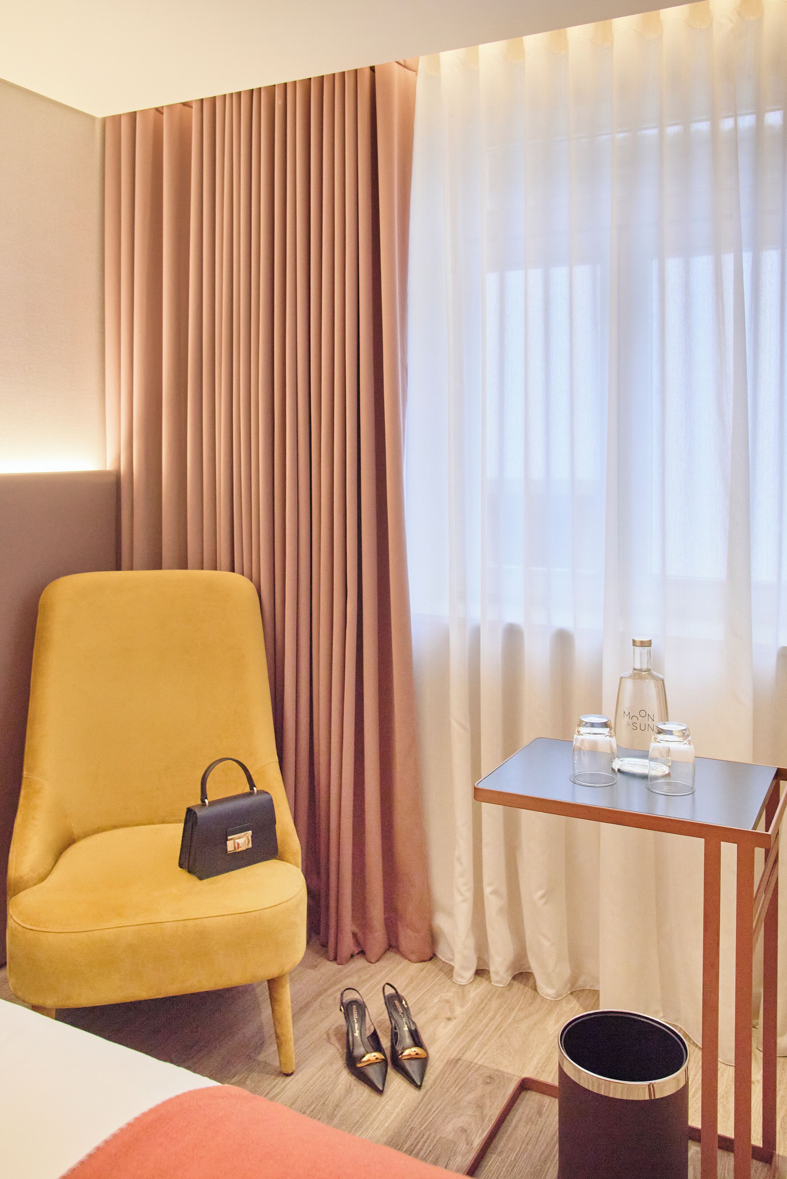 Habitación de hotel con un sillón amarillo, una mesita con una botella y vasos, y un par de zapatos en el suelo.
