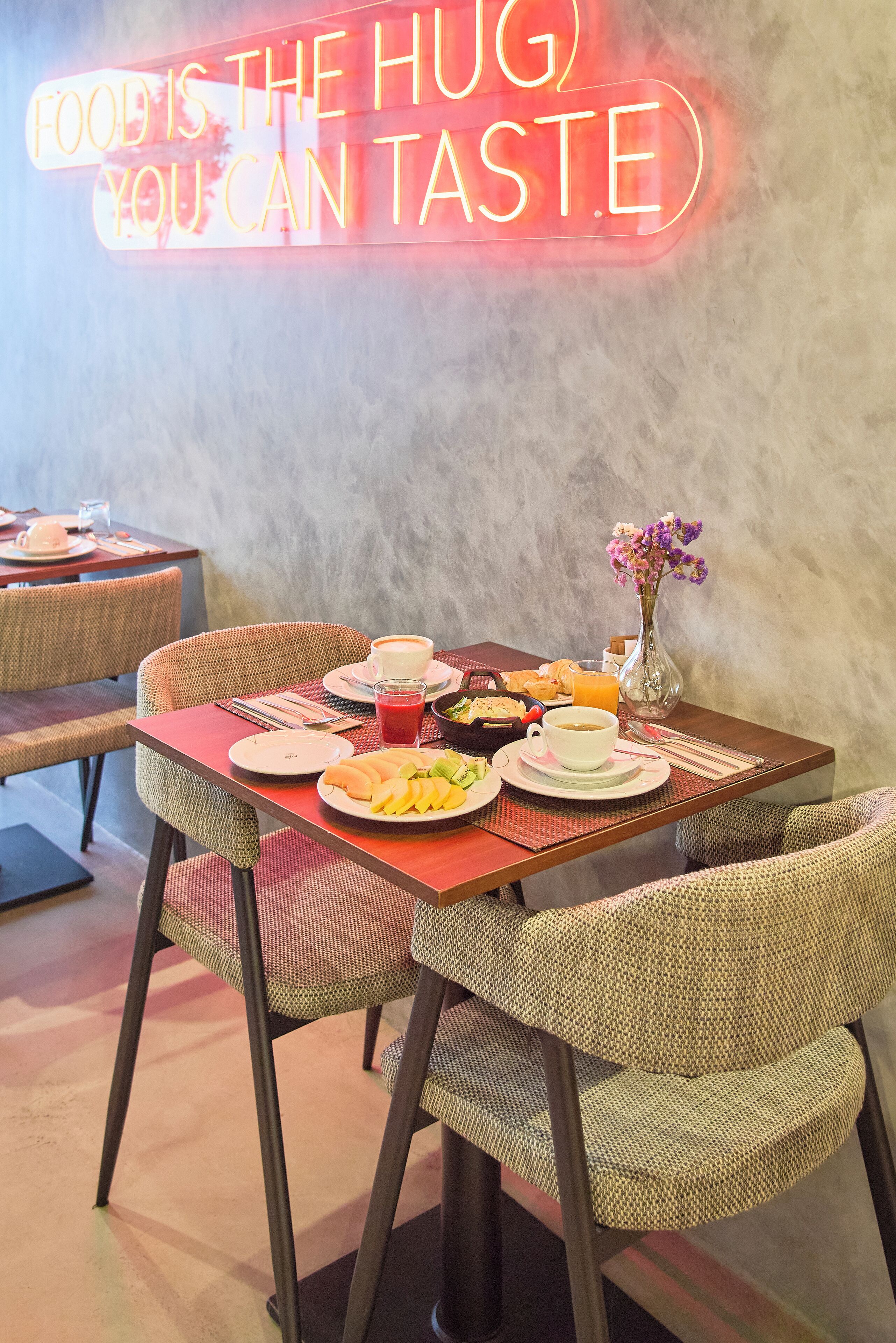 Un restaurante con letrero de neón, sillas de mimbre, una mesa puesta con platos, un jarrón de flores y un menú.