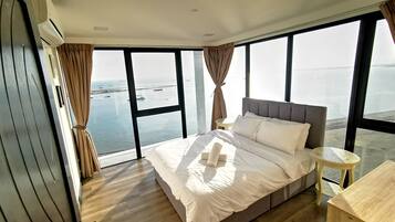 A bedroom with a large bed, a small bedside table, and a view of the sea through large windows.