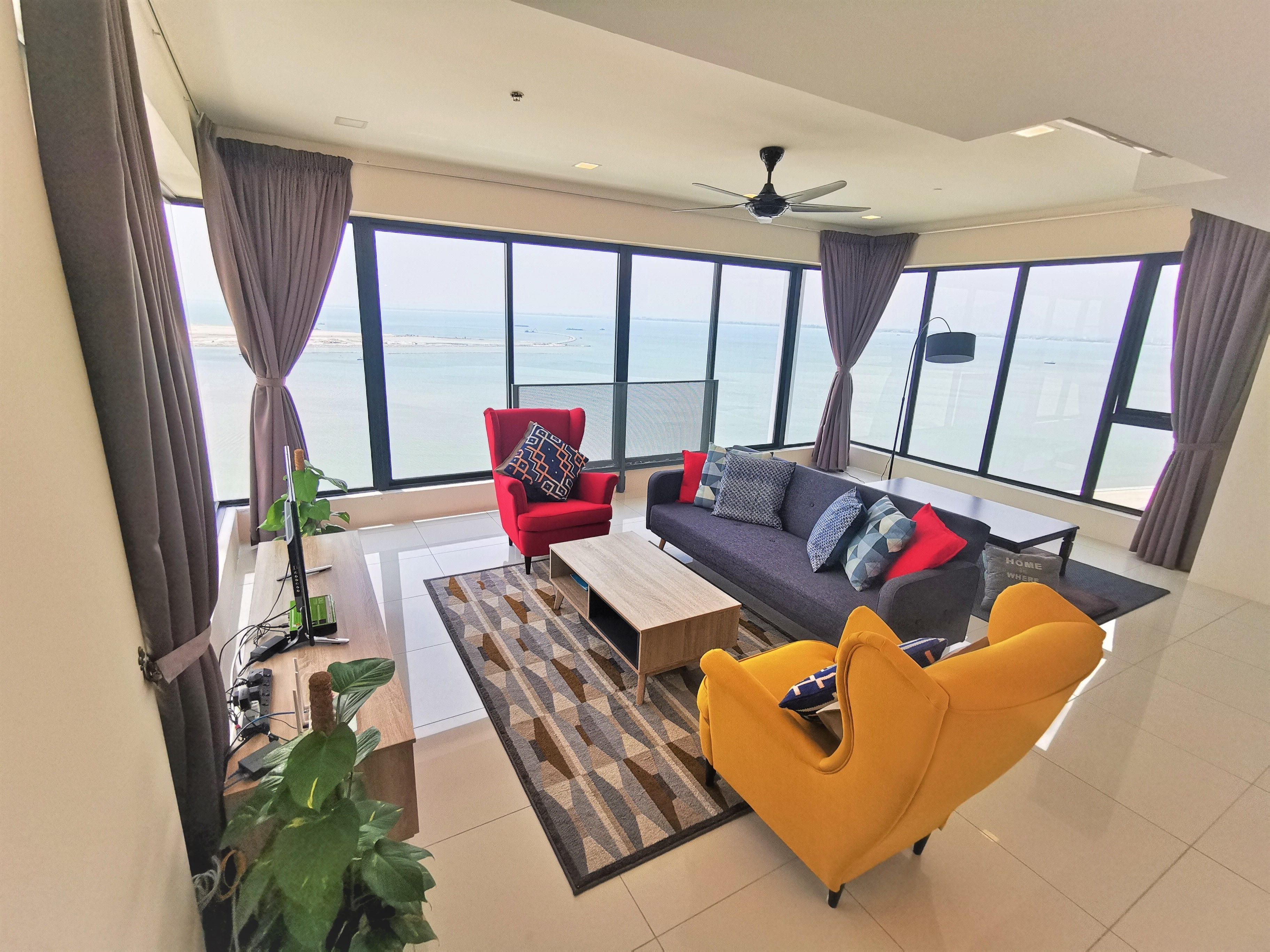 Een ruime woonkamer met een groot raam met uitzicht op zee, een rode fauteuil, een gele fauteuil, een donkere bank en een plafondventilator.