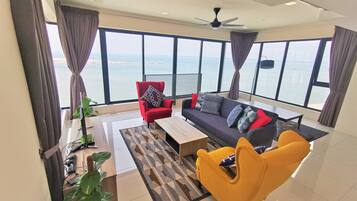 Een ruime woonkamer met een groot raam met uitzicht op zee, een rode fauteuil, een gele fauteuil, een donkere bank en een plafondventilator.