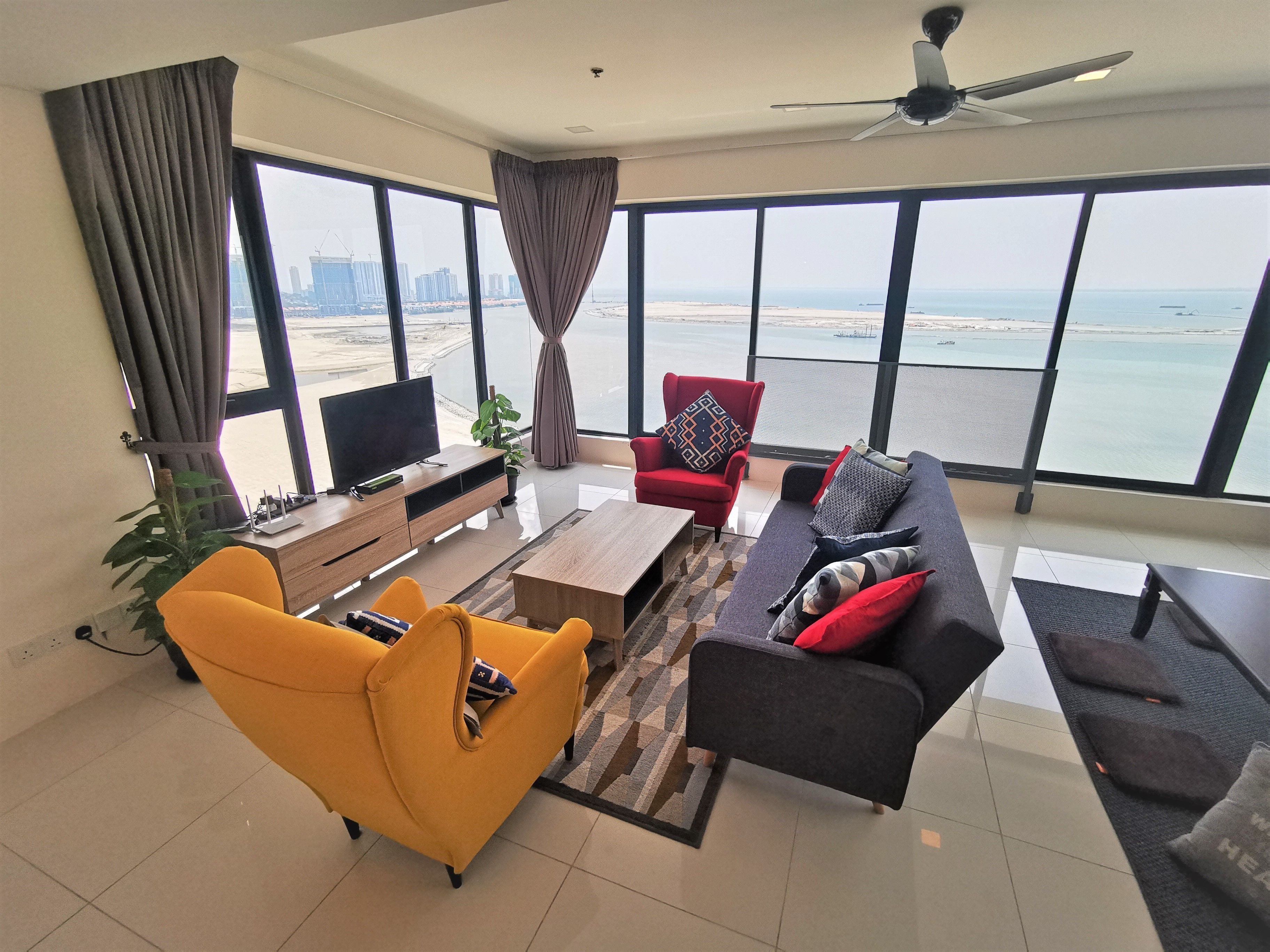 A modern living room with a sofa, armchair, and TV, large windows with a view of the sea and buildings, and a ceiling fan.