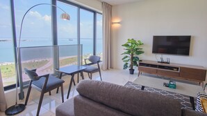 A modern living room with a large window, a sofa, a TV, and a dining area. - Family Suite Sunrise Gurney Penang (George Town)