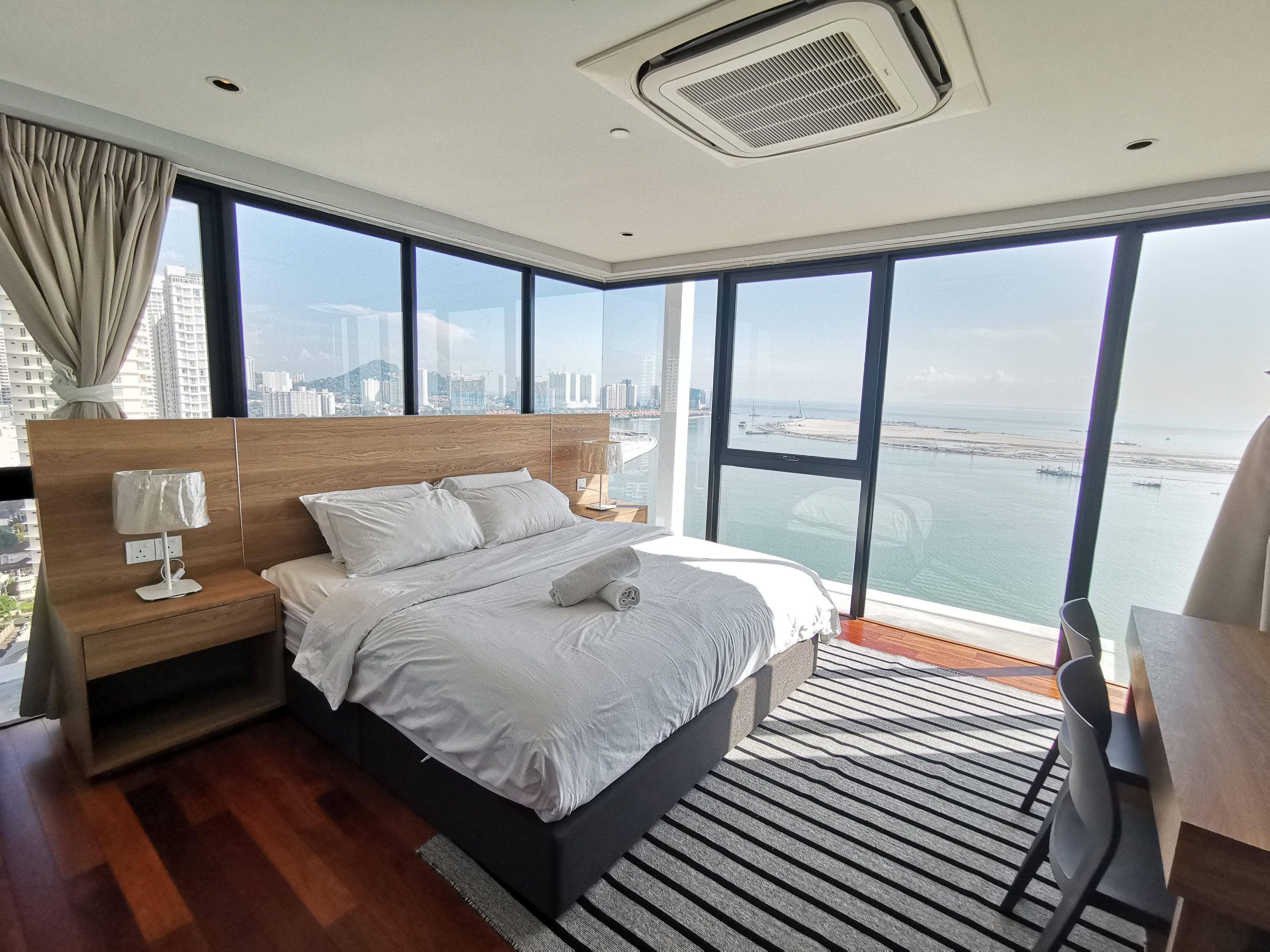 Een moderne hotelkamer met een groot bed, een houten hoofdbord, een nachtkastje met een lamp en uitzicht op de stad en zee door grote ramen.