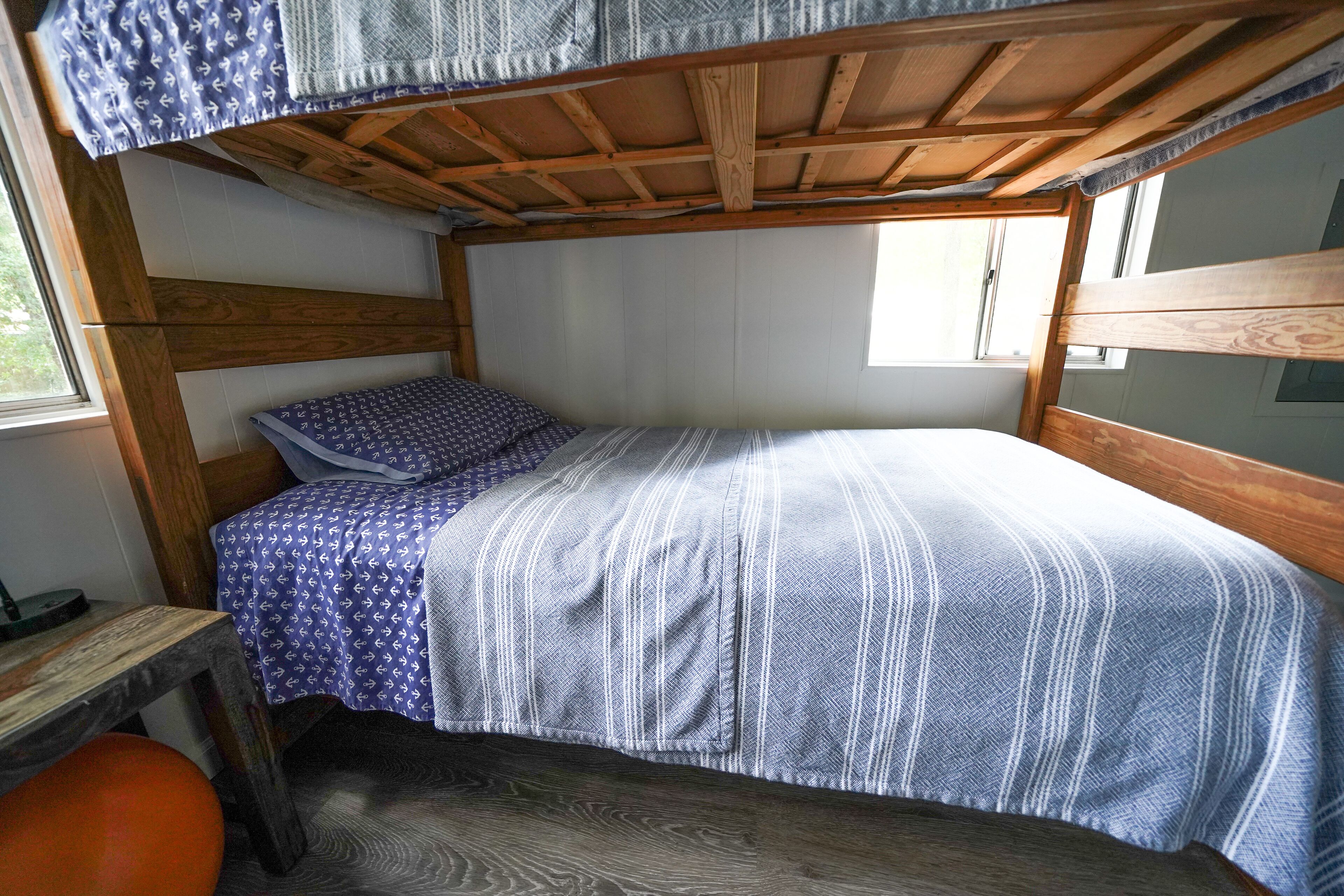 A small, single-bed room with a wooden bunk bed structure, a window, and a small table with a chair.