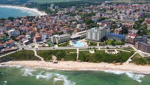 An aerial view of a coastal town with a central swimming pool surrounded by buildings, a beach, and a road. - Hotel Perla Beach Luxury (Primorsko)