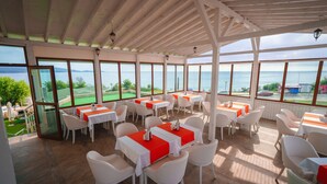 A restaurant with white tables and chairs, large windows with a view of the sea, and a wooden ceiling. - Hotel Perla Beach Luxury (Primorsko)