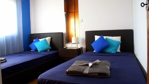 A hotel room with two beds, each with a blue bedspread and matching pillows. There is a nightstand with a lamp between the beds. - Panoramic Blue (Limassol)
