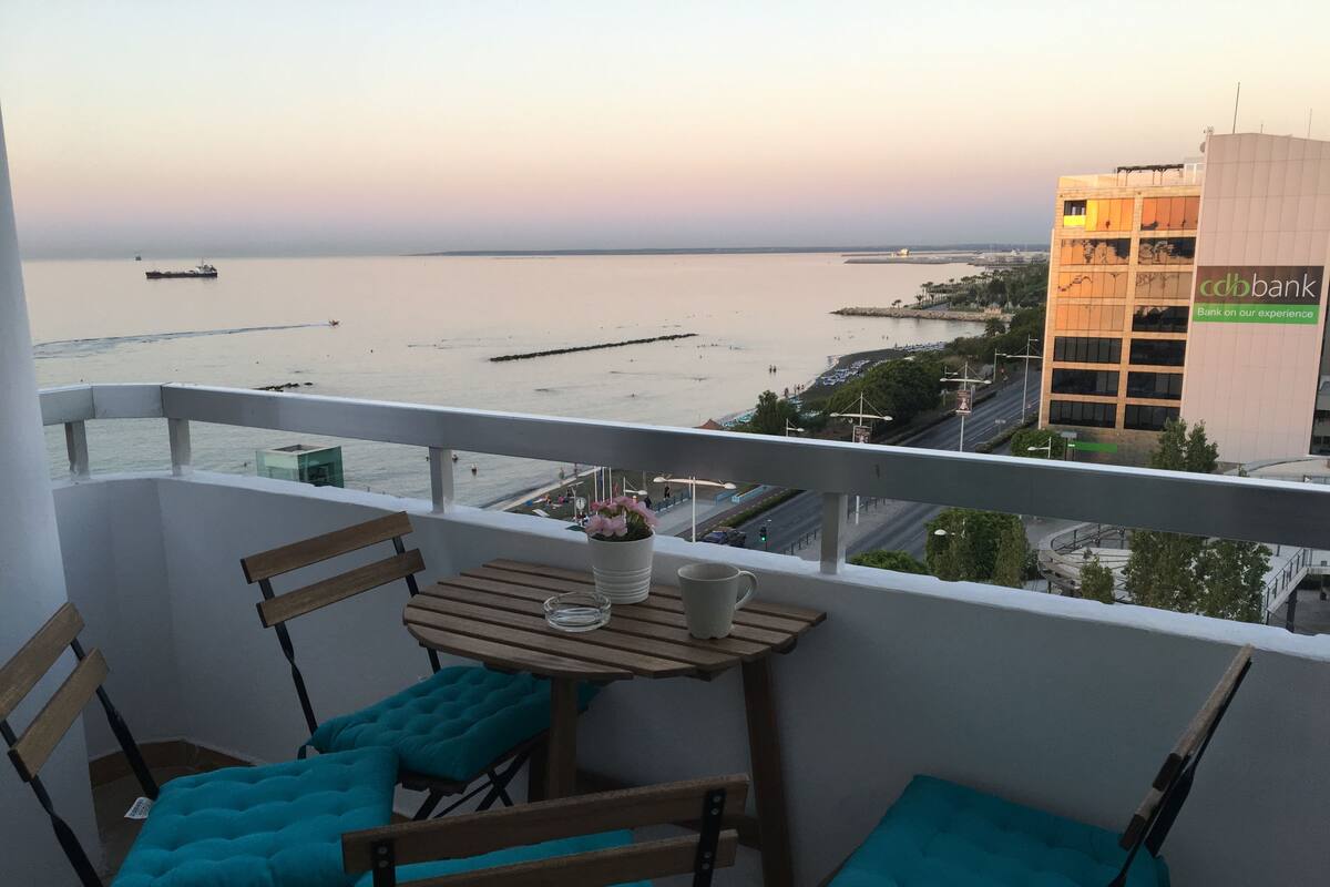 A balcony with a table and chairs, overlooking a beach and a cityscape.