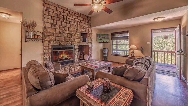 A living room with a stone fireplace, a ceiling fan, and a large window. - Deer Crossing: Soak, Stargaze, Relax (Ruidoso)