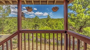 A wooden balcony with railings, hanging flower baskets, and a view of a forested hillside. - Deer Crossing: Soak, Stargaze, Relax (Ruidoso)