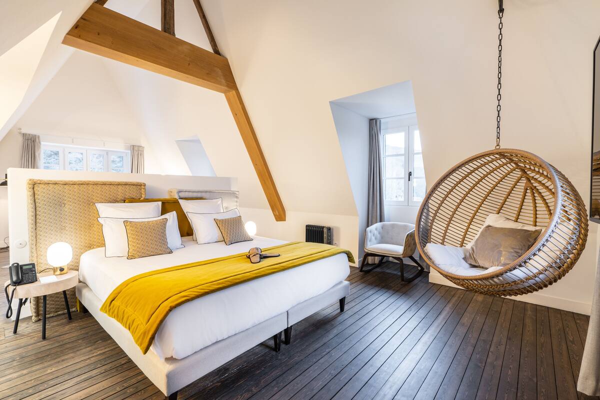 A modern bedroom with a large bed, a hanging chair, and a small table with a lamp.