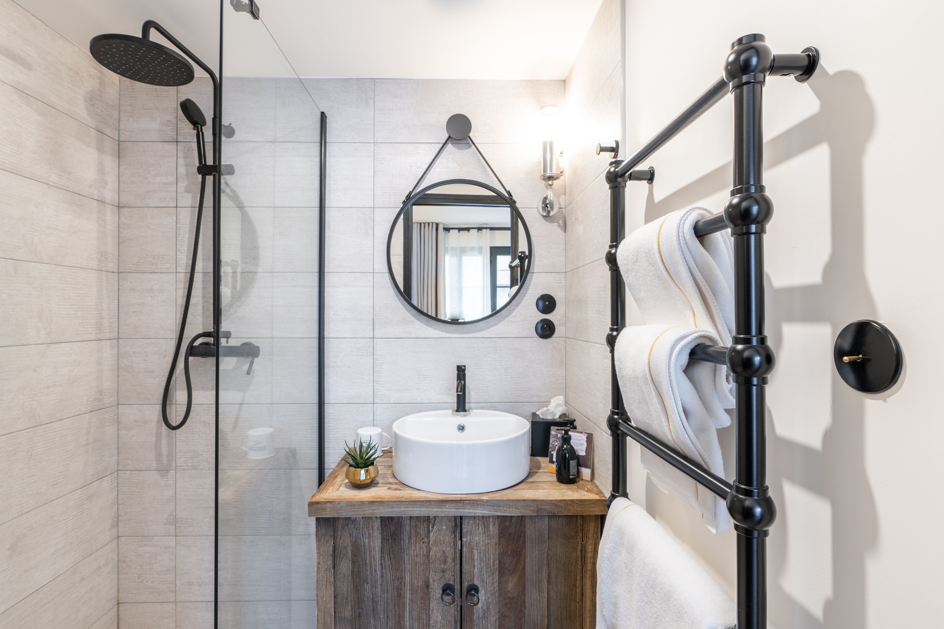 Un baño moderno con tocador de madera, espejo redondo, lavamanos blanco, ducha y toallero.