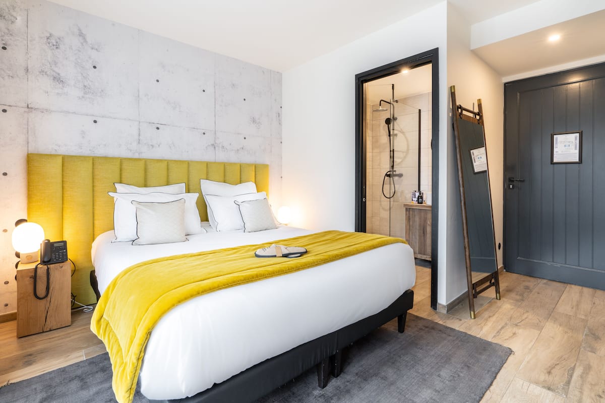 A modern bedroom with a large bed, a wooden nightstand with a lamp, and a bathroom with a shower and a wooden ladder.