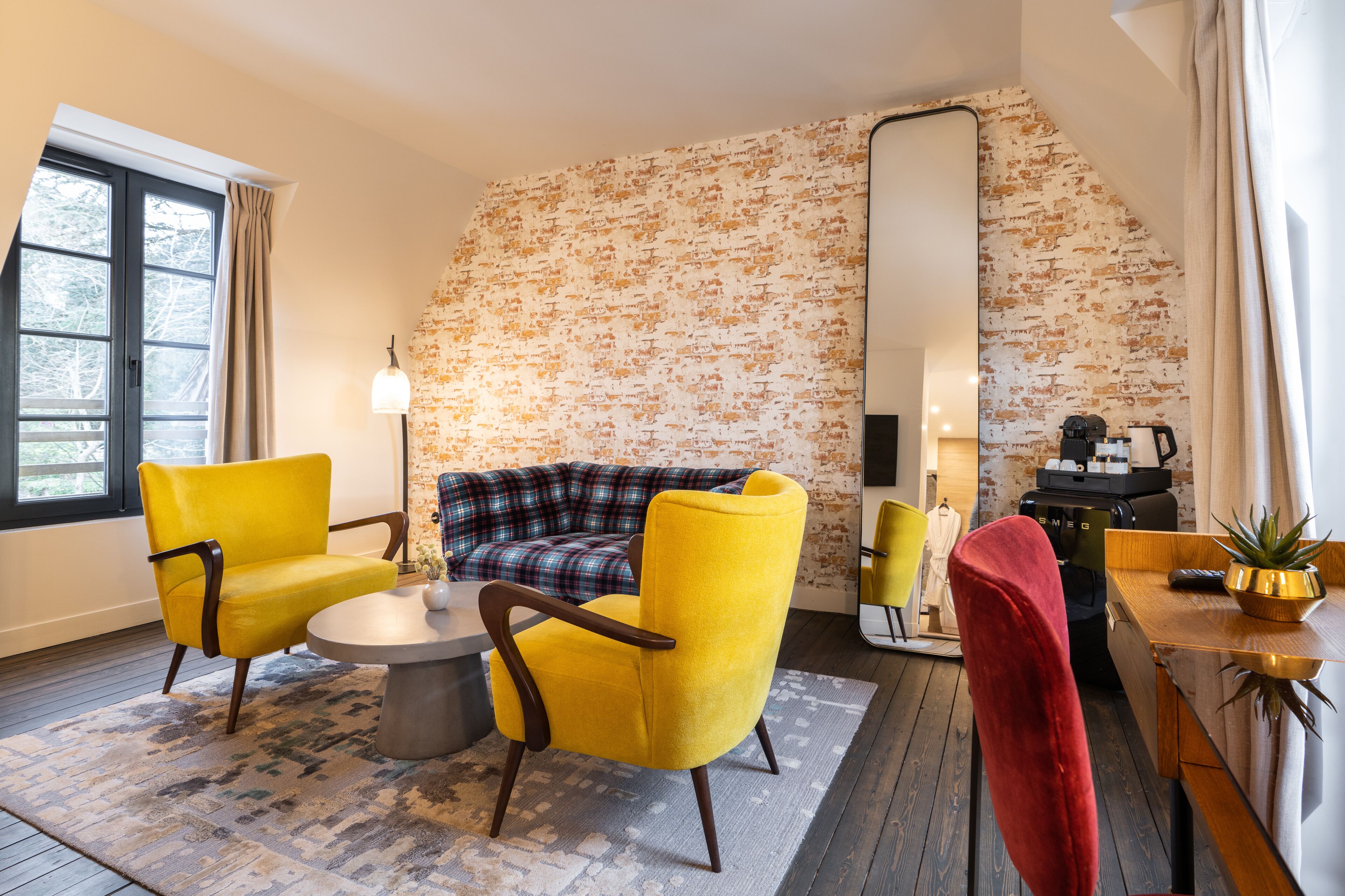 A room with a sofa, two yellow armchairs, a red chair, a coffee machine, and a large mirror.