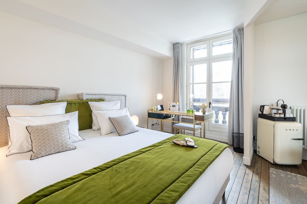 A hotel room with a large bed, a desk, a chair, a radiator, and a window with curtains.