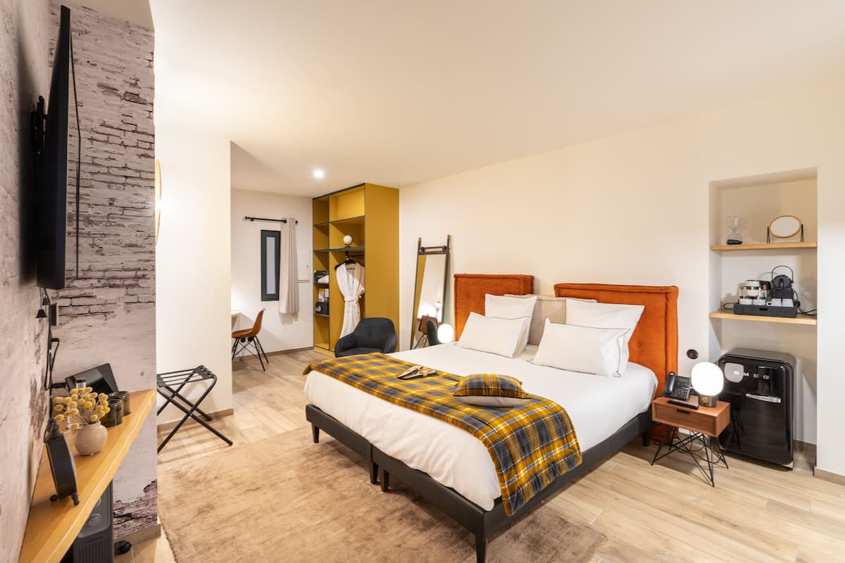 A modern hotel room with a large bed, a flat-screen TV mounted on a brick wall, a small desk with a lamp, and a wardrobe.