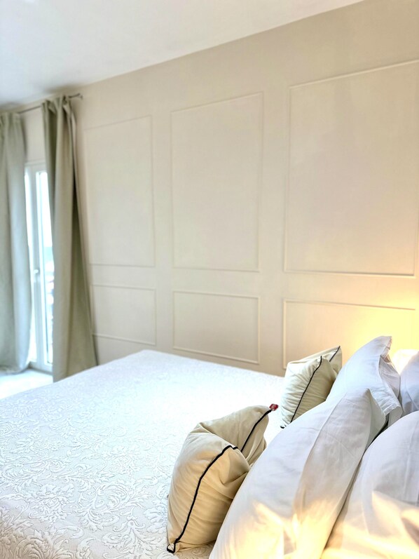 A neatly made bed with white linens and beige pillows, a plain wall with paneling, and a window with curtains. - B&B Eyexei Domus (Agrigento)