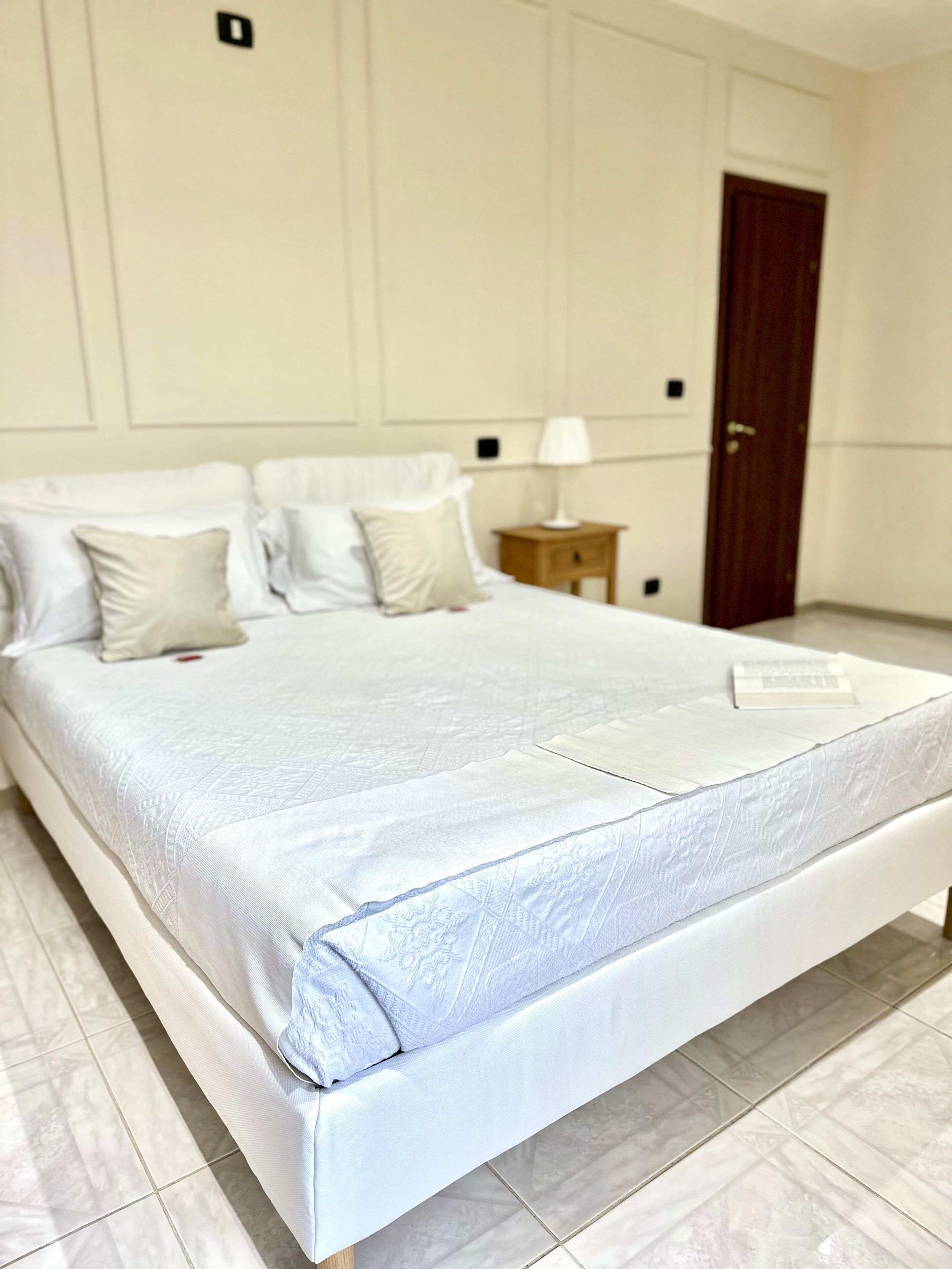 A neatly made bed with a white bedspread and pillows, a wooden nightstand with a lamp, and a wooden door in a room with white walls and tiled flooring.
