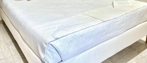 A neatly made bed with a white bedspread and pillows, a wooden nightstand with a lamp, and a wooden door in a room with white walls and tiled flooring.