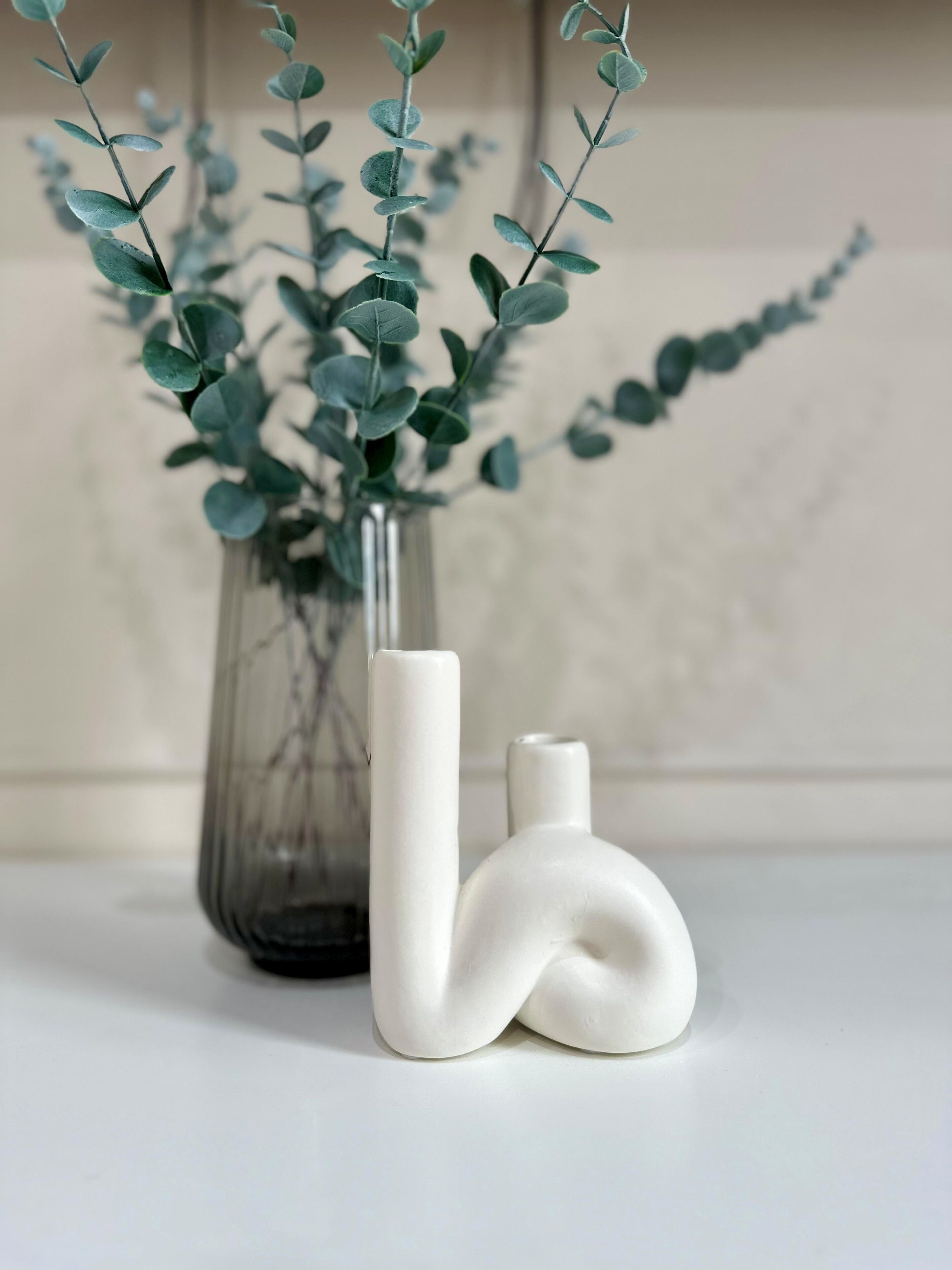A clear glass vase with a plant, a white ceramic sculpture, and a white ceramic vase on a white surface.