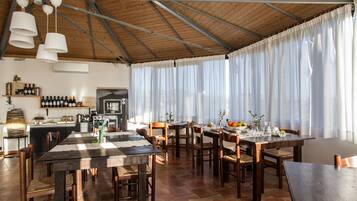 Un comedor amplio con mesas y sillas de madera, una cocina al fondo y ventanas grandes con cortinas traslúcidas.