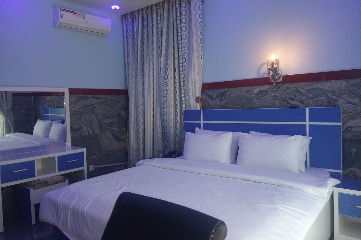 A hotel room with a bed, a nightstand, a mirror, a window with curtains, and an air conditioning unit.