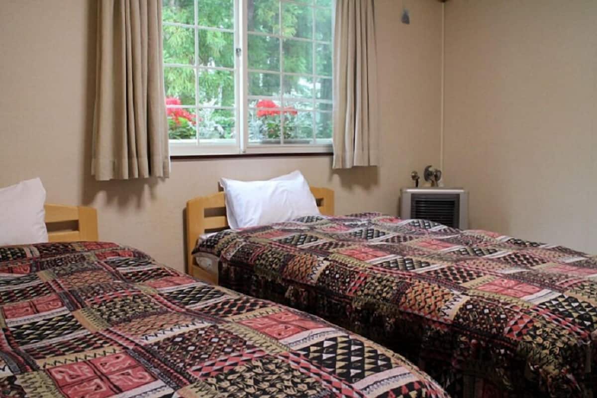 A room with two beds, each with patterned quilts, a window with curtains, and a radiator.