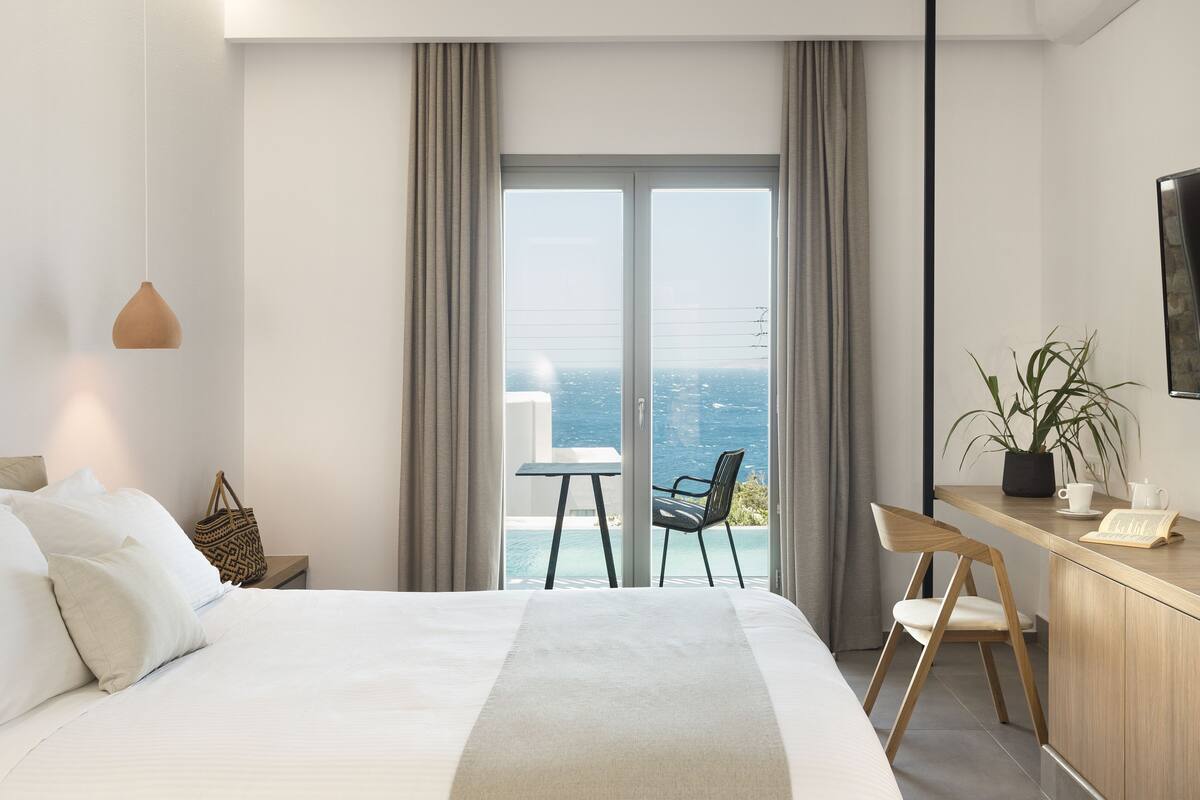 Ett modernt hotellrum med en stor säng, ett skrivbord med en stol, utsikt över havet och en krukväxt.