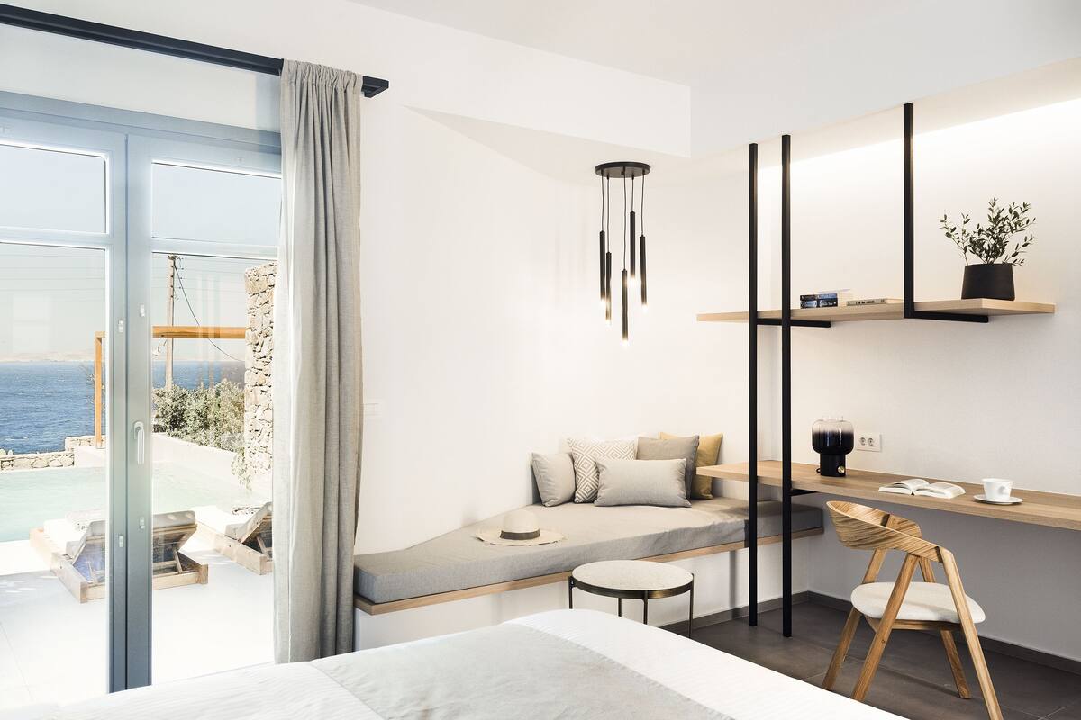 Ett modernt hotellrum med en säng, en sittgrupp med utsikt över havet, ett skrivbord med en växt och en hängande lampa.