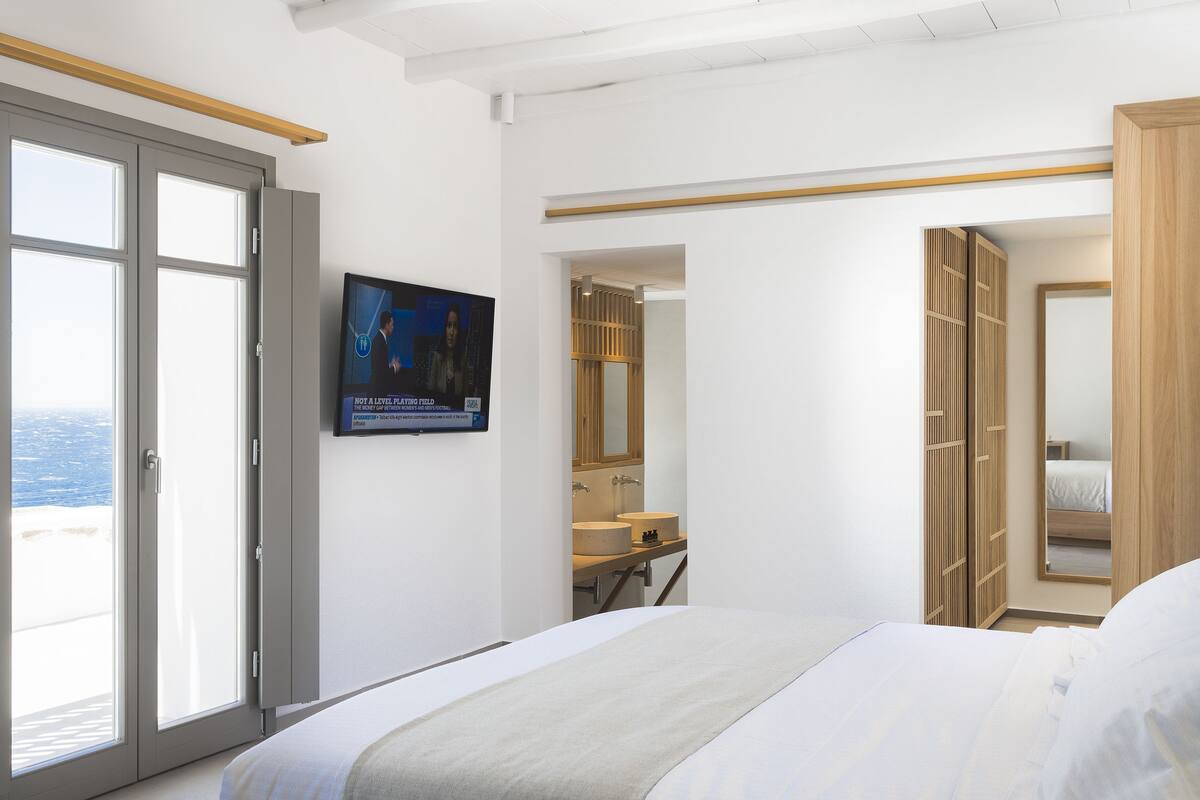 Ett hotellrum med en säng, en tv som är monterad på väggen, en garderob i trä och ett badrum med handfat och spegel.
