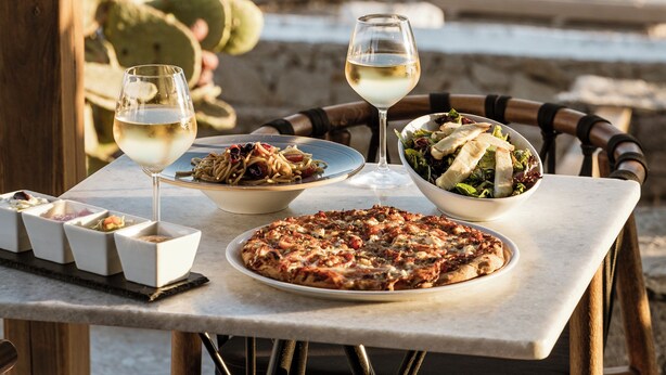 Een tafel met pizza, salade en wijnglazen, met uitzicht op de kust.