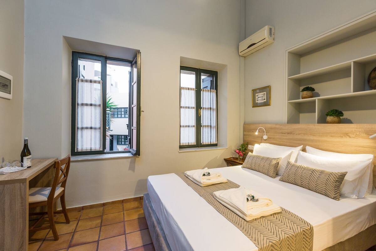 A hotel room with a bed, a desk, two windows, a shelf with decorative items, and a wall-mounted air conditioner.