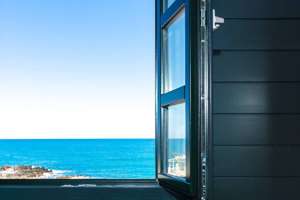 A room with a view of the ocean through an open window, a dark-colored wall, and a cushion with a geometric pattern.