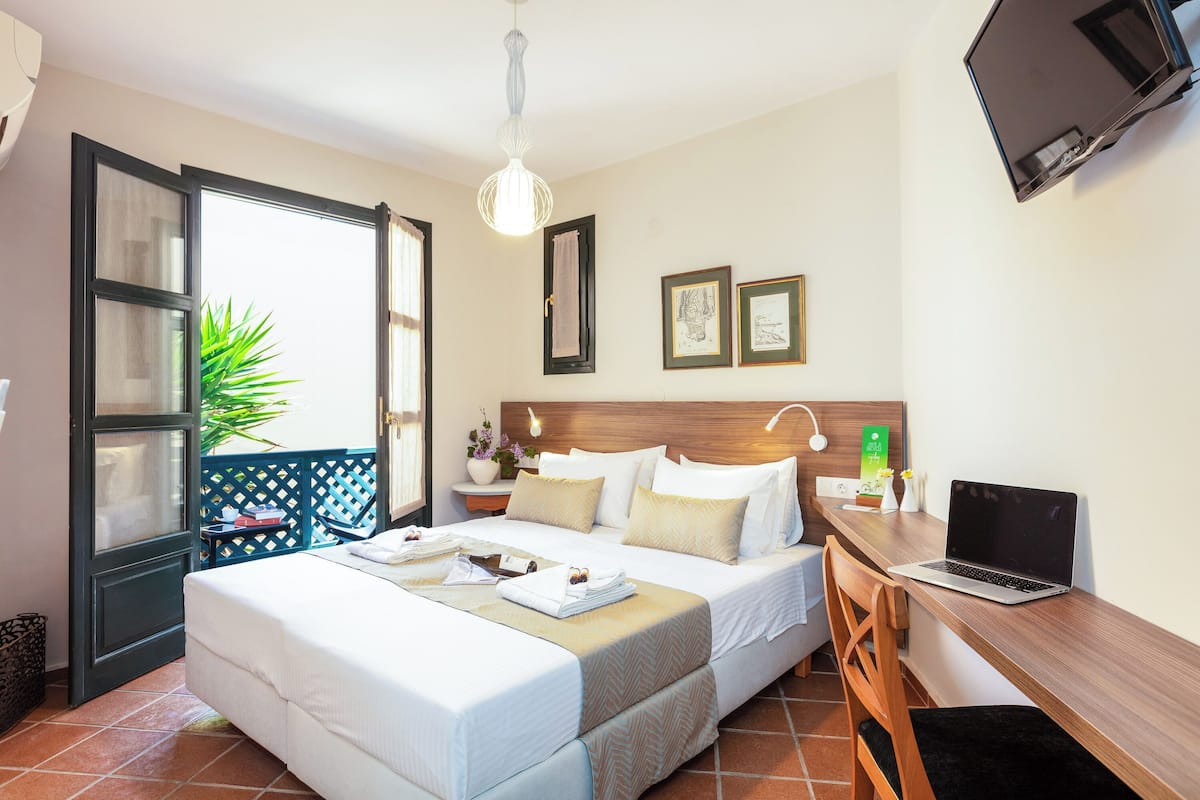 A hotel room with a bed, a desk with a laptop, a TV, a window with curtains, and a plant.