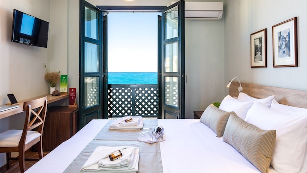 A hotel room with a bed, a desk, a chair, a TV, and a view of the sea through an open door. - Ink Hotels House of Europe (Rethymno)