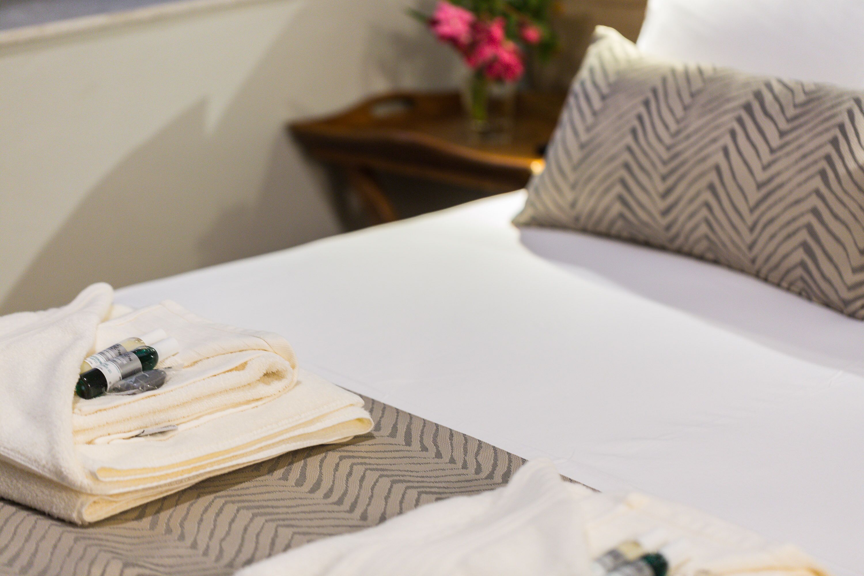 Una cama bien hecha con una toalla doblada y una botella de loción, un cojín estampado y una pequeña mesa con un florero al fondo.