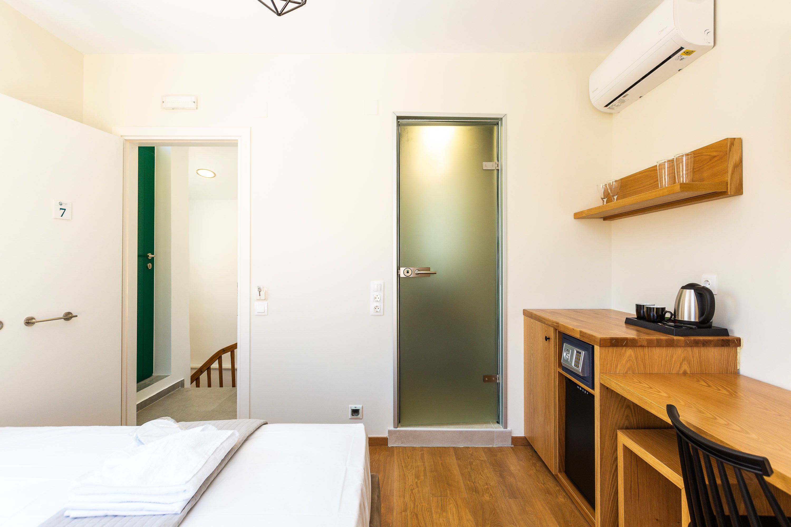 A compact hotel room with a bed, a small desk with a chair, a coffee maker, and a door with frosted glass.