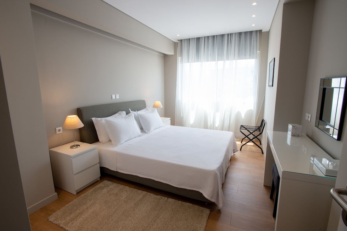 Quarto de hotel moderno com uma cama grande, mesas de cabeceira, uma TV e uma escrivaninha de vidro.