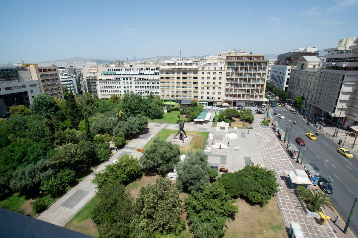 Paisagem urbana com um edifício imponente, uma praça central com uma escultura e uma rua movimentada com veículos.