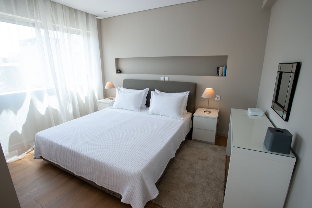 Quarto de hotel bem arrumado, com uma cama grande, mesas de cabeceira, uma escrivaninha e um espelho na parede.