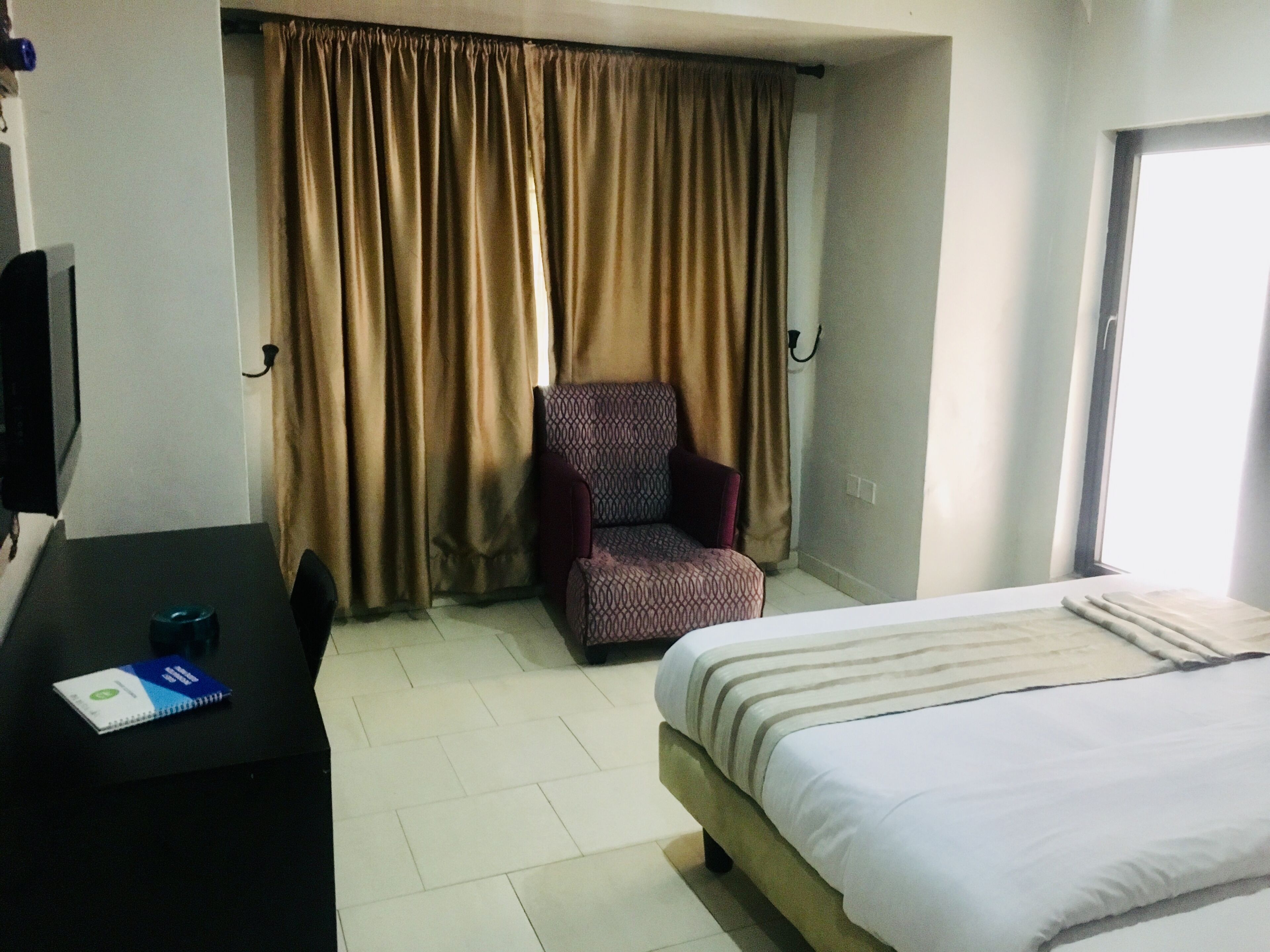 Habitación de hotel con cama, escritorio con libreta, silla y ventana con cortinas.