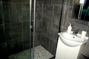 A modern bathroom with dark tiles, a glass shower enclosure, and a white sink with toiletries. - Blue Home (Limassol)
