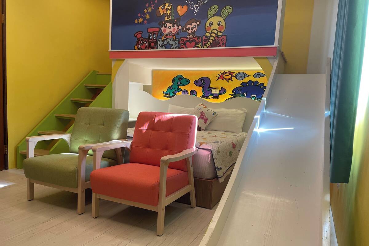 A children's playroom with a bed, two chairs, a staircase, and colorful wall decorations.