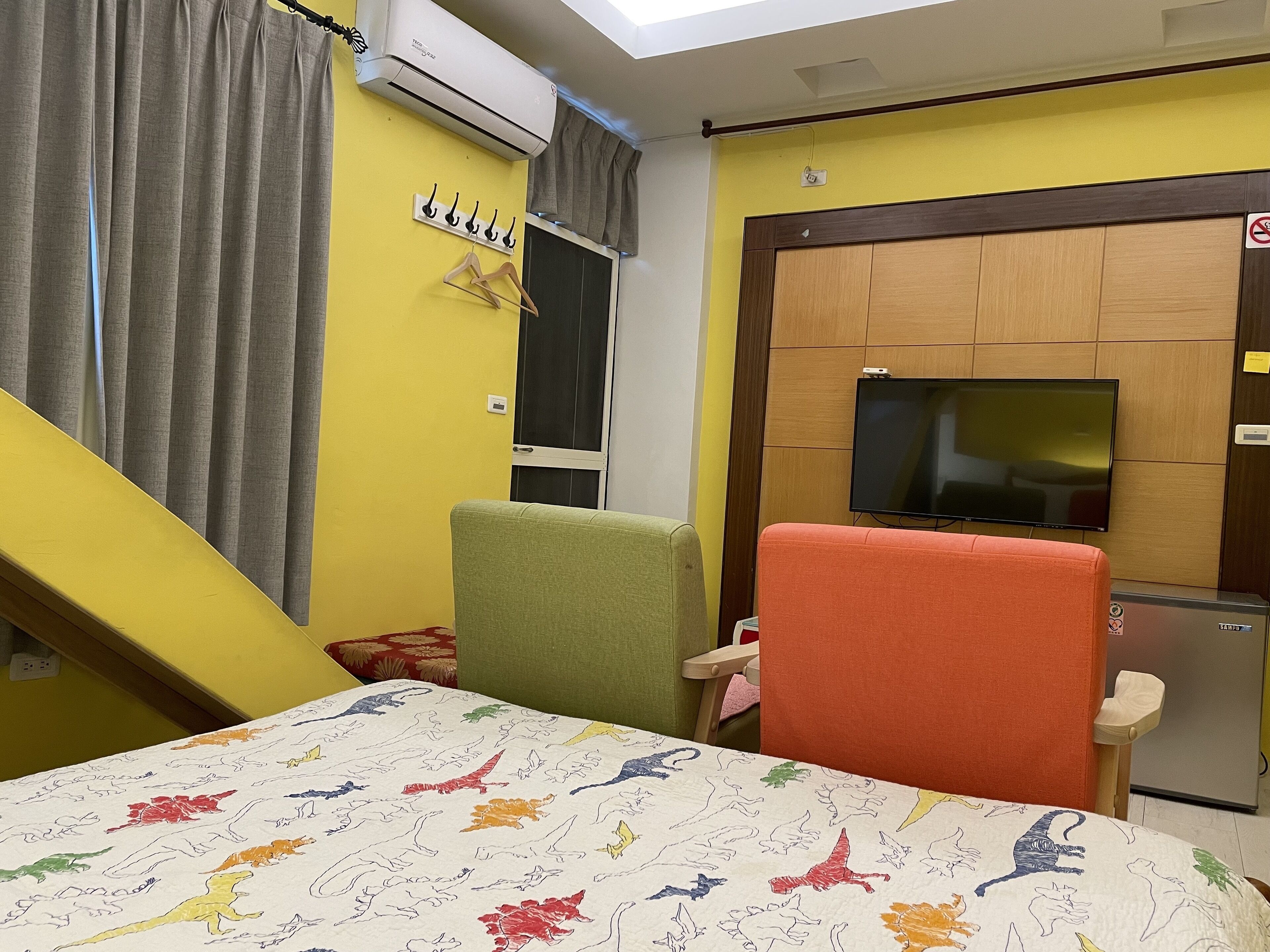 A room with a yellow wall, a mounted TV, a sofa, a chair, and a bed with a dinosaur-patterned bedspread.
