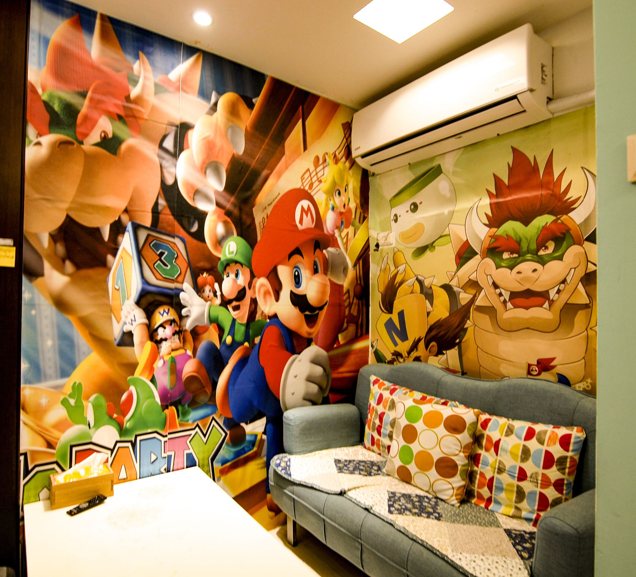 A room with a Mario-themed mural, a gray sofa with colorful pillows, and an air conditioning unit on the wall.