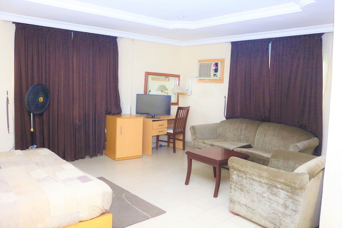 A hotel room with a bed, a desk with a computer, a sofa, a chair, and a fan.