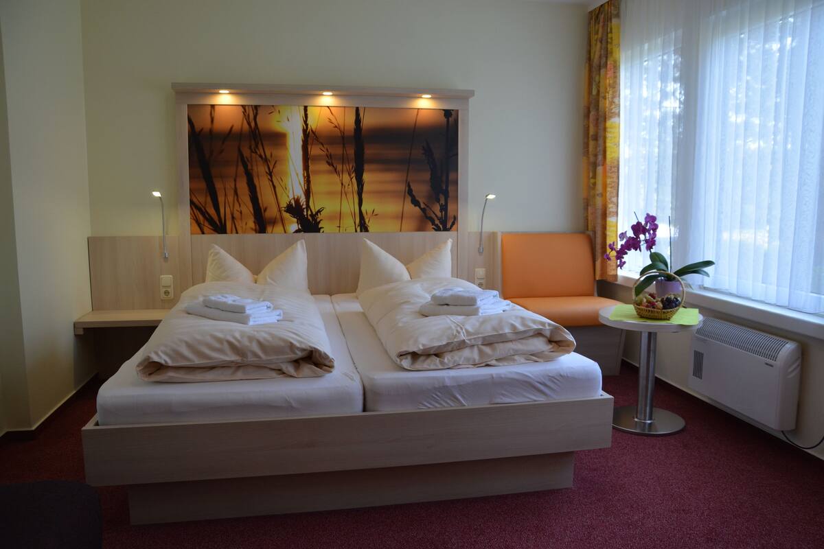 A double bed with white linens, a small round table with a basket of fruit, and a window with curtains.