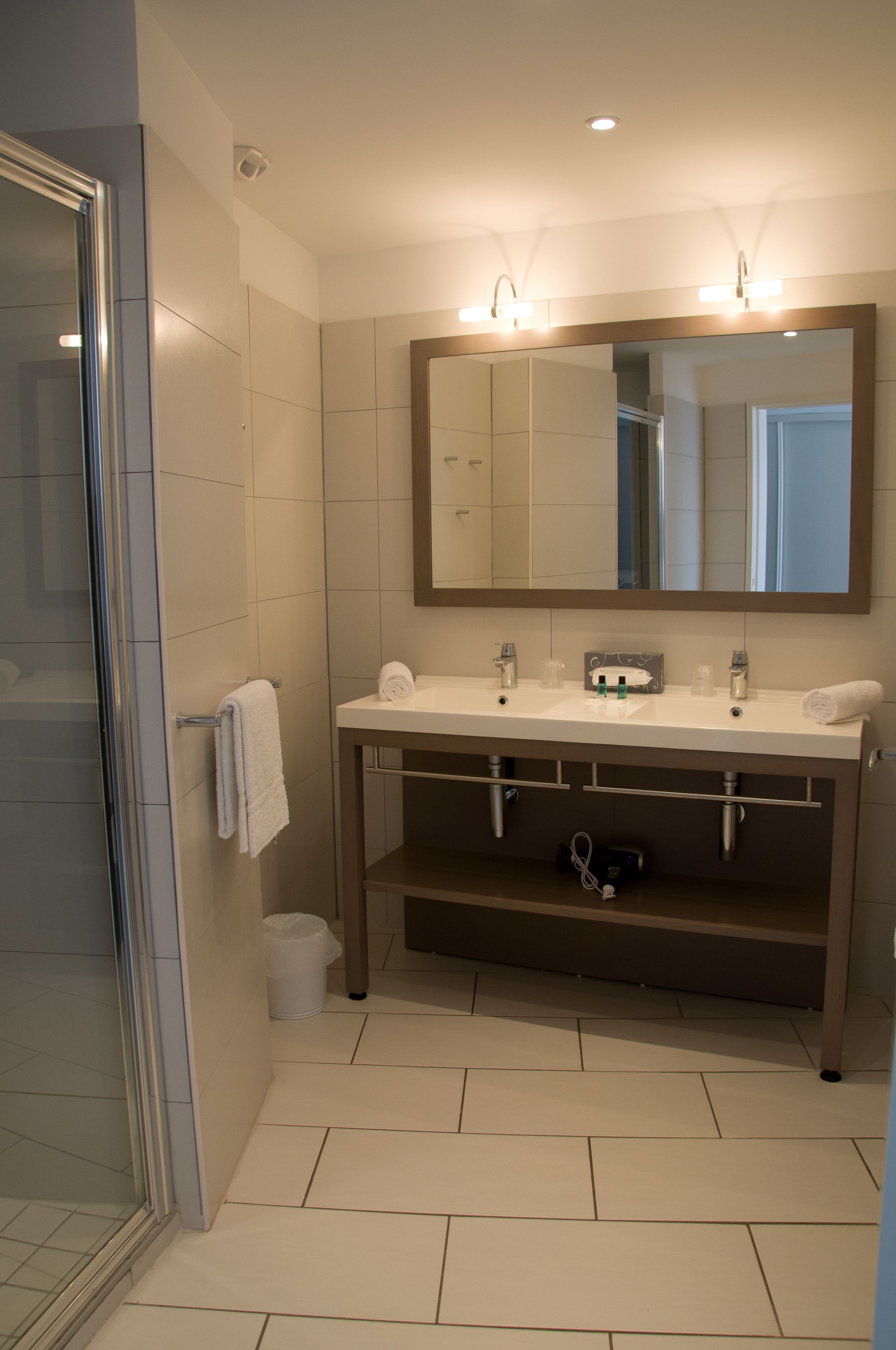 Une salle de bain moderne avec une douche à l’italienne en verre, un lavabo blanc avec deux miroirs et un porte-serviettes.