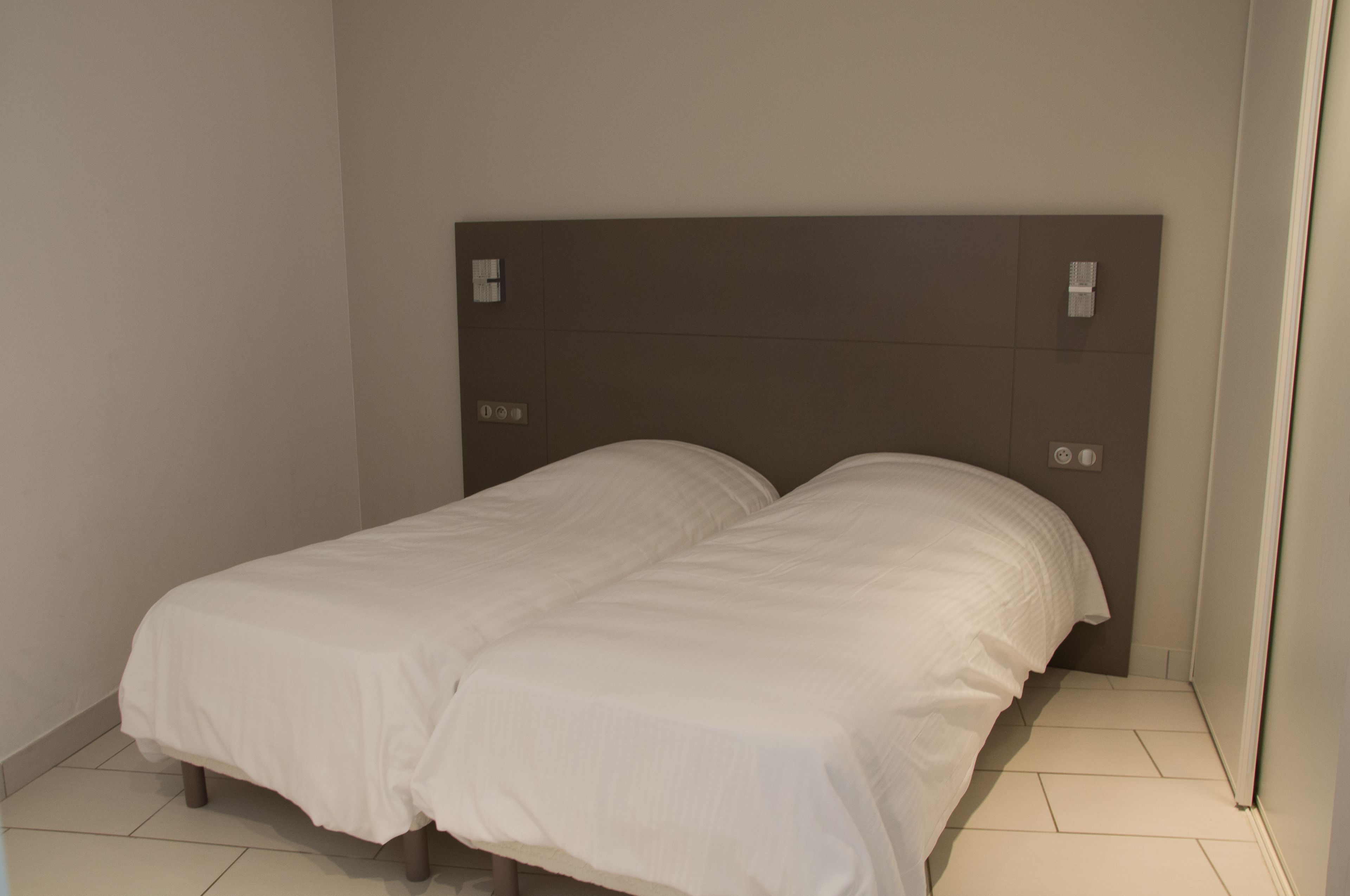 Cama doble con cabecera sencilla y ropa de cama blanca en una habitación pequeña con suelo de baldosas.